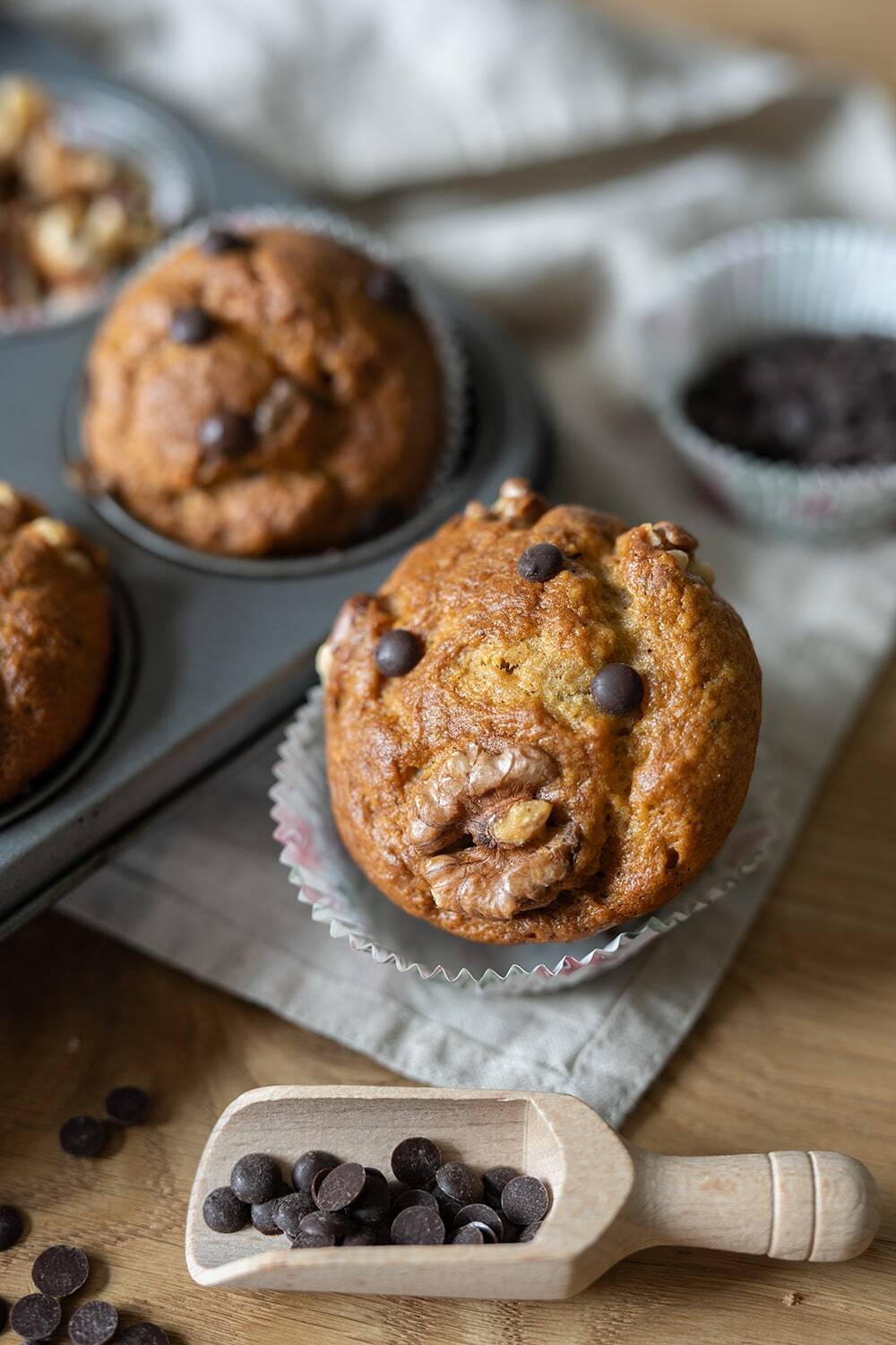 Ein Schokoladenchip-Muffin liegt in einem Papiereinsatz auf einer Holzfläche, weitere Bananenmuffins liegen in einer Backform in der Nähe. Im Vordergrund befindet sich ein mit Schokoladenstückchen gefüllter Holzlöffel neben einem grauen Tuch.