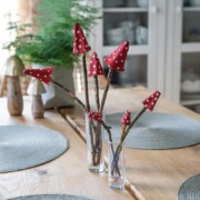Zwei Gläser auf einem Holztisch halten Stäbchen mit roten, gepunkteten Pilzhüten - eine charmante Bastelidee für die Herbstdeko. Weitere Pilzdekorationen und Tischsets sind zu sehen, perfekt als einfache Bastelideen für saisonales Basteln.
