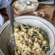 Auf einem Holztisch steht eine Schüssel mit cremigem Risotto mit Wirsing und kleinen Stücken von Pancetta. Ein Löffel ruht in der Schüssel, geriebener Käse liegt in einer Schale daneben, und eine grün-blaue Serviette suggeriert köstliche gesunde Rezepte.
