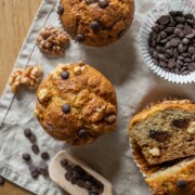 Draufsicht auf Muffins mit Schokoladenstückchen auf einem beigen Tuch, mit Schokoladenstückchen, Walnüssen und einem Holzlöffel in der Nähe. Ein Muffin ist in der Mitte durchgeschnitten, so dass man die Schokoladensplitter im Inneren sieht - die perfekte Inspiration für neue Muffinrezepte wie Bananenmuffins ohne Zucker.