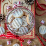Ein mit Weihnachtskeksen gefülltes Glas, das mit einer roten Schleife verziert ist, steht auf einem dekorativen Buch. Süßigkeiten, Tannenzapfen und rote Bänder sind auf der Holzoberfläche verstreut und schaffen eine gemütliche Weihnachtsszene.