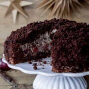 Ein Schokokuchen mit einer cremigen Füllung und Kirschen, ausgestellt auf einem weißen Kuchenständer. Ein Stück ist ausgeschnitten, und im Hintergrund befinden sich dekorative Papiersterne. Frische Kirschen stehen neben diesem festlichen Weihnachtskuchen.