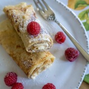 Zwei gerollte Crêpes, inspiriert von einem klassischen Rezept Pfannkuchen, werden mit Sahne gefüllt, mit Puderzucker bestäubt und mit frischen Himbeeren belegt. Weitere Himbeeren werden mit einer Gabel auf einen weißen Teller gestreut.