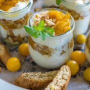 Gläser mit geschichtetem Dessert mit Schlagsahne, Pfirsichen und zerbröselten Keksen, garniert mit Minzblättern. Gelbe Kirschtomaten und Biscotti umgeben die Gläser auf einem runden Serviertablett.