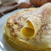 Ein Stapel dünner, goldfarbener Pfannkuchen liegt auf einem weißen Teller, darauf ein aufgerollter Crêpe. Im unscharfen Hintergrund sind Eier und ein Behälter mit Mehl zu sehen, die auf die klassischen Rezept-Pfannkuchen-Zutaten hinweisen.