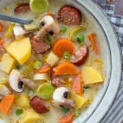 Eine Schüssel mit cremigem Kartoffeleintopf mit geschnittener Wurst, Kartoffeln, Karotten, Champignons und Lauch, garniert mit grünen Zwiebeln. Ein Löffel liegt in der Schüssel und Brot ist an der Seite zu sehen.