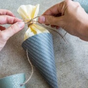 Hände binden Bindfäden um eine kegelförmige Schultüte, die in gemustertes Papier und hellgelbes Seidenpapier eingewickelt ist, perfekt als Einschulungsdeko, mit anderen ähnlichen Kegeln in der Nähe auf einer grauen Fläche.