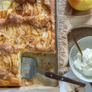 Ein Tablett mit in Scheiben geschnittenem Apfelkuchenblech mit Mandelblättchen und einem Tortenheber steht neben einem ganzen Apfel, einer kleinen Schüssel mit Schlagsahne und herbstlichen Dekorationen auf einer Holzfläche.