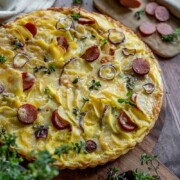 Eine herzhafte Kartoffelquiche mit geschmolzenem Käse, in Scheiben geschnittenen gelben Kartoffeln, roten Zwiebelringen und Wurstscheiben, garniert mit frischen Kräutern auf einem Holzbrett, mit zusätzlicher Wurst und Kräutern in der Nähe - perfekt für jeden Liebhaber von Tarte-Rezepten.