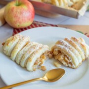 Ein weißer Teller mit zwei mit Puderzucker bestäubten Apfeltaschen, einem goldenen Löffel und einem frischen Apfel, wobei im Hintergrund weiteres Gebäck zu sehen ist.