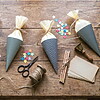 Vier handgefertigte Schultüten mit gelbem Krepppapier, umgeben von buntem Sternkonfetti, einer Schere, Schnur, Bleistiften, Karten und einem Wachssiegelstempel, sorgen auf einem rustikalen Holztisch für eine bezaubernde Einschulungsdekoration.