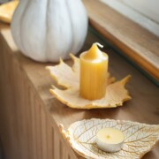 Eine gelbe Kerze und ein Teelicht stehen auf blattförmigen Keramiktellern aus Modelliermasse DIY, perfekt für Basteln mit Kindern im Herbst, auf einer Holzfläche am Fenster mit einem weißen Zierkürbis im Hintergrund.