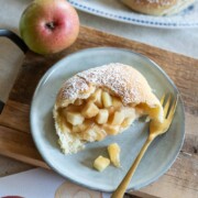 Ein Teller mit aufgeschnittenen, mit Apfelstückchen gefüllten und mit Puderzucker bestäubten Buchteln, daneben eine goldene Gabel. Im Hintergrund sind ein Apfel und weitere Hefegebäckstücke zu sehen, daneben ein Schneidebrett und eine Apfelillustration.