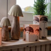 Auf einer Fensterbank stehen dekorative Lampen in Pilzform und quadratische Upcycling-Laternen mit Pilzmotiven, die alle sanft leuchten. Neben den selbstgebastelten Lampen stehen auch grüne Topfpflanzen, die perfekt für Bastelfreunde sind.