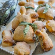 Ostergebäck in Form von Hasengebäck, hergestellt aus Quark-Öl-Teig-Hasen und mit Zucker bestreut, wird auf einem Teller angerichtet. Einige sind mit grün-weiß karierten Bändern verschnürt, im Hintergrund stehen ein Drahtkorb mit verzierten Eiern und eine Serviette.