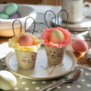 Zwei dekorierte Eierbecher mit Hasengesichtern und Drahtohren halten bunte Eier auf einem Teller - perfekt für Osterbrunch Ideen. Weitere bunte Eier und eine Schale mit Eiern stehen im Hintergrund auf einem Holztisch mit gepunktetem Tuch, Löffel und Tasse.