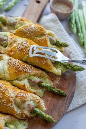 Goldene, in Blätterteig gewickelte Spargelstangen, ein köstliches Spargelrezept, werden mit Sesam bestreut und auf einem Holzbrett serviert. Mit einem Metallspatel wird ein Stück herausgehoben - perfektes Fingerfood für jede Zusammenkunft. Frischer Spargel und eine Schale mit Sesamkörnern runden das Bild ab.