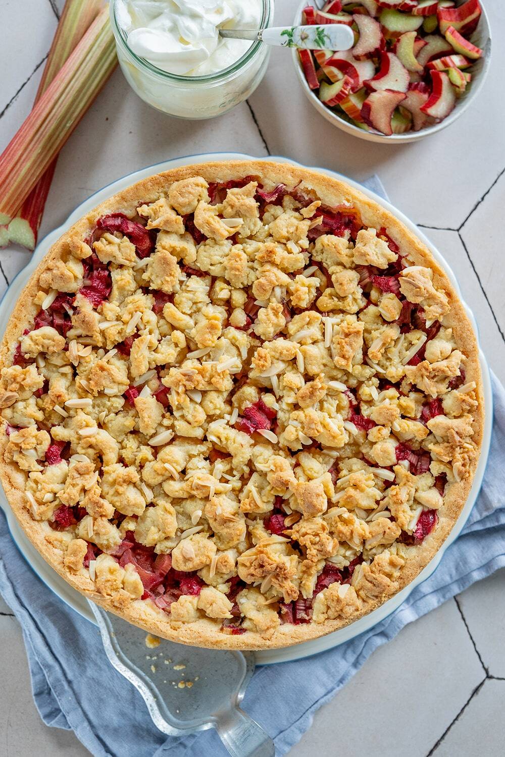 Ein runder Erdbeer-Rhabarber-Streuselkuchen, inspiriert von einem klassischen Rhabarberkuchenrezept, liegt mit einem goldenen Belag auf einem blauen Tuch. Daneben liegen frische Rhabarberstangen, geschnittenes Obst und ein Glas Schlagsahne.