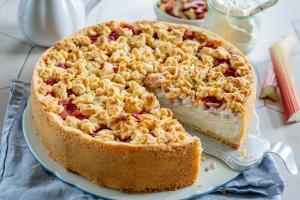 Ein runder Rhabarberkuchen mit goldenem, bröckeligem Belag und cremiger Füllung liegt auf einem weißen Teller, eine Scheibe fehlt. Umgeben von Rhabarberstangen und einer Schale mit Sahne ist dieser Kuchen perfekt für alle, die ein authentisches Rhabarberkuchenrezept suchen.