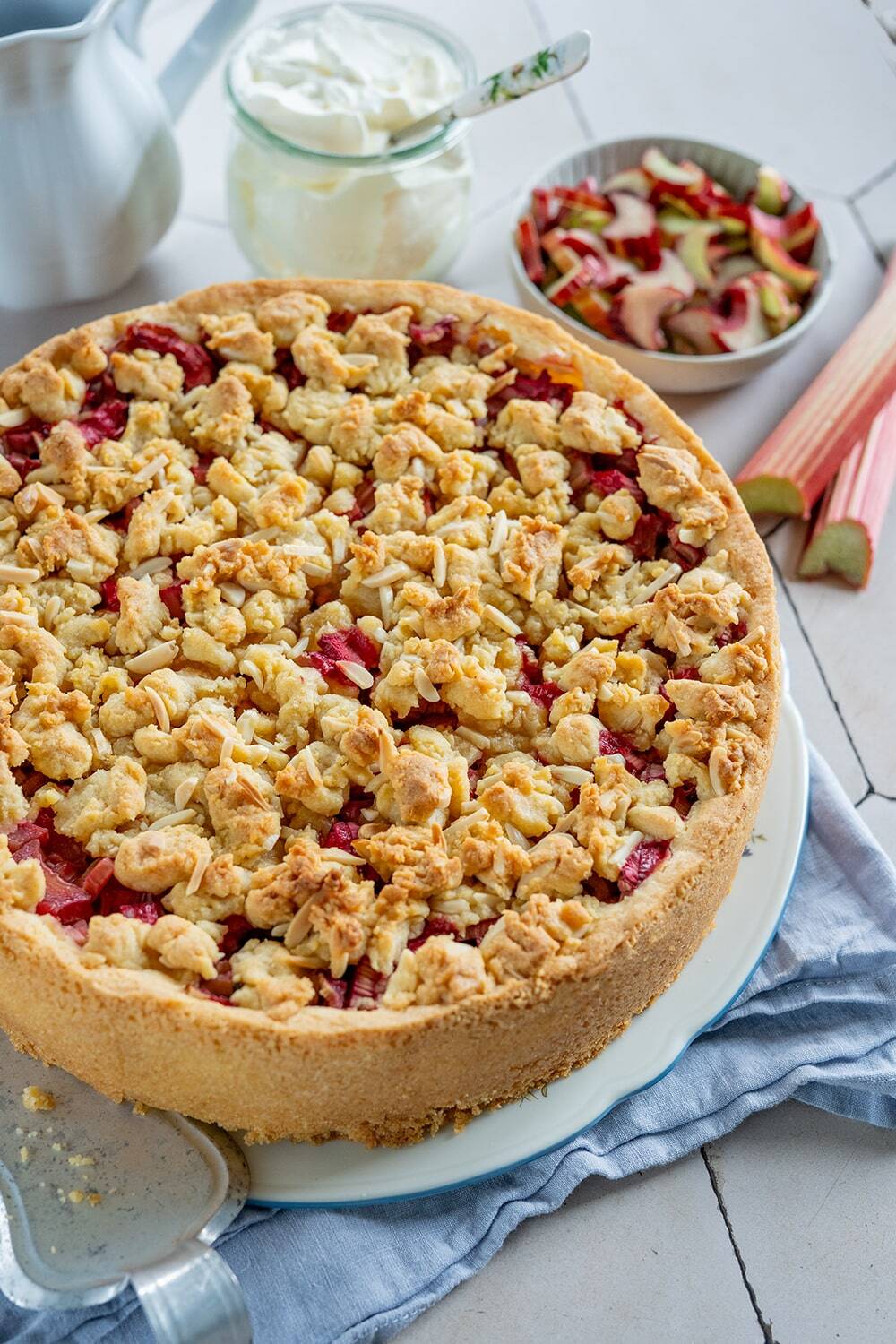 Auf einem weißen Teller liegt ein runder Rhabarberkuchen mit Pudding, der mit goldenen Streuseln belegt ist. Daneben stehen auf einem weißen Tisch eine Schüssel mit gehacktem Rhabarber, ein Glas Sahne und ein Krug.