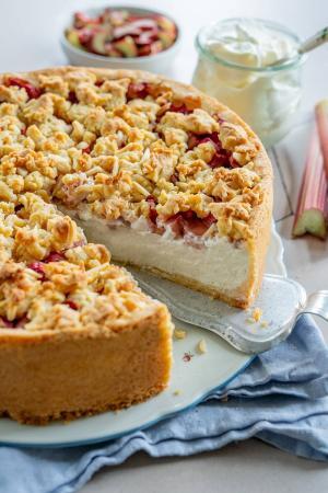 Eine Nahaufnahme eines Rhabarberkuchens mit Pudding mit krümeligem Belag und einem Stück, das serviert wird. Der Kuchen hat eine dicke Kruste, eine cremige Füllung und Fruchtstückchen. Im Hintergrund stehen Gläser mit Sahne und gehackten Früchten auf einer hellen Fläche.