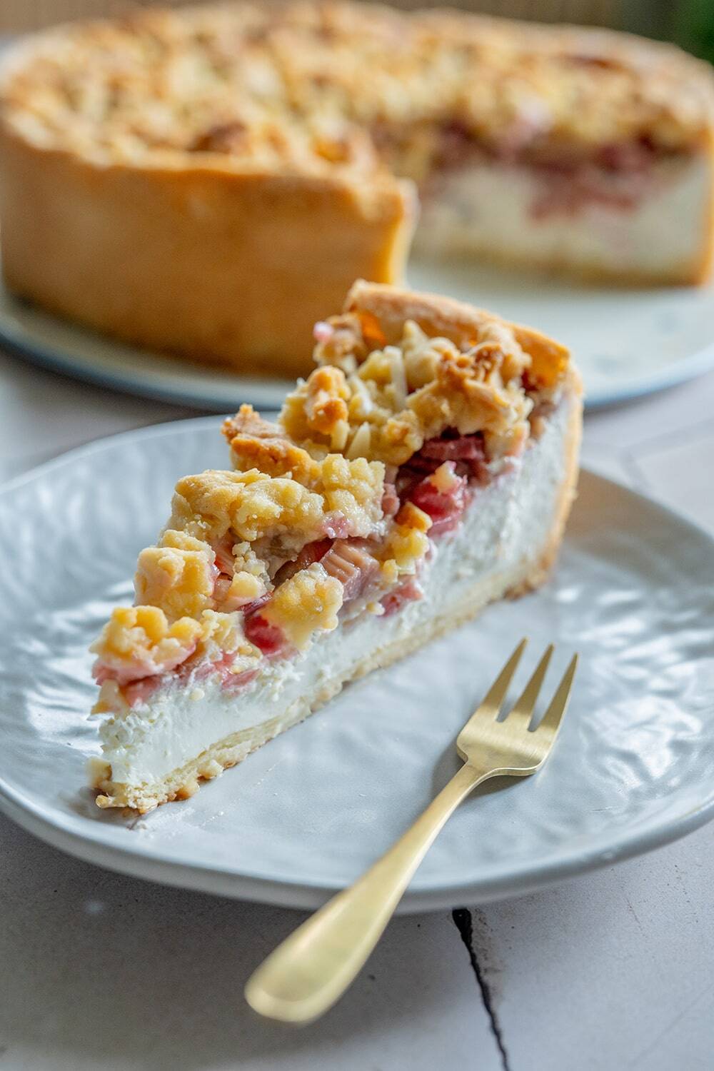 Ein Stück Rhabarberkuchen mit Pudding und sichtbaren Fruchtstücken liegt auf einem weißen Teller mit einer goldenen Gabel. Der Rest des Kuchens befindet sich im Hintergrund auf einem separaten Teller.