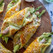 Herzhafter Blätterteig, gefüllt mit Spargel und Schinken, bestreut mit Sesamsamen, präsentiert auf einem Holzbrett. Serviert wird dieses köstliche Fingerfood mit einem Schälchen Sesam und einem Teller mit grünem Dip daneben.