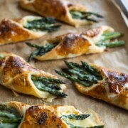 Goldene Blätterteighüllen, gefüllt mit grünen Spargelstangen und geschmolzenem Käse, angerichtet auf einem mit Pergamentpapier ausgelegten Backblech - diese Spargel im Blätterteigmantel ergeben die perfekte knusprige und frisch gebackene Spargelvorspeise.