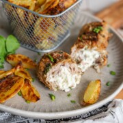 Ein Teller mit gerösteten Kartoffelecken und einer Bifteki-Fleischrolle, gefüllt mit cremiger Käsefüllung, garniert mit gehackten Kräutern. Frische Petersilie und weitere Kartoffelspalten in einem Metallkorb vervollständigen das Gericht.