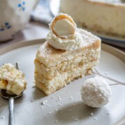 Ein Stück cremiger Raffaello-Tiramisu-Kuchen mit weißem Schokoladenkonfekt liegt auf einem Teller neben einem runden Leckerbissen und einem Löffel Kuchen - perfekt für alle, die Lust auf Kuchen ohne Backen haben, wobei ein ganzer Kuchen und ein Becher im Hintergrund verschwimmen.