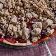 Nahaufnahme eines Zwetschgenkuchens mit Streusel, mit goldenem, krümeligem Belag und sichtbarer roter Fruchtfüllung, auf einer Glasplatte mit einem Servierlöffel auf einem rosa strukturierten Tuch.