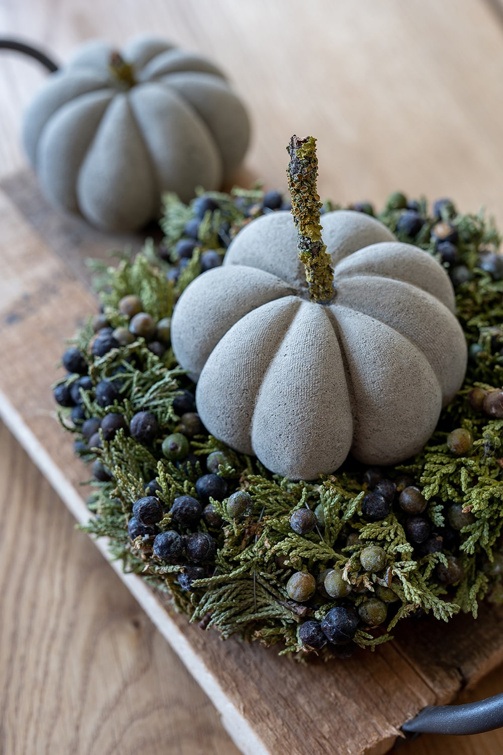 Ein Stoffkürbis mit grünem, strukturiertem Stiel sitzt auf einem Kranz aus Grünzeug und kleinen Beeren, perfekt als Herbstdeko. Im Hintergrund ist ein weiterer Stoffkürbis zu sehen, der eine charmante Bastelidee für Ihr Zuhause darstellt.
