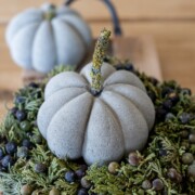 Ein Stoffkürbis mit strukturiertem Stiel sitzt auf einem Kranz aus grünem Laub und dunklen Beeren auf einer hölzernen Oberfläche - eine charmante Bastelidee von Herbstdeko. Ein anderer ähnlicher Stoffkürbis ist im Hintergrund verschwommen.