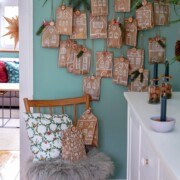 Ein an der Wand befestigter Adventskalender aus braunen Papiertüten, die als Häuser dekoriert sind, hängt an Tannenzweigen. Darunter steht ein Holzstuhl mit einem Kissen und einer Pelzdecke, auf dem weitere Füllung für Adventskalender in einem gemütlichen, festlichen Raum stehen.