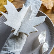 Auf einem grauen Teller auf einem Holztisch liegt eine Stoffserviette mit einem Serviettenring aus weißem Filz mit Ahornblättern - perfekt für Basteln mit Filz. Getrocknete Blätter liegen verstreut auf dem Tisch und sorgen für ein herbstliches Ambiente.