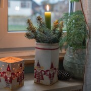 Eine grüne Kerze mit Tannenzweigen in einem festlichen Weihnachtsmann-Halter steht auf der Fensterbank neben einem leuchtenden Laternenbasteln und einer Vase mit Tannenzweigen. Ein Tannenzapfen und ein Vorhang verleihen dieser gemütlichen Weihnachtsdeko Basteln Charme.