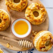 Ein Holztablett mit gebackenen Donuts, die von Bienenstich inspiriert sind, mit karamellisierten Mandeln bestreut und einer kleinen Schale mit Honig und einem Honiglöffel umgeben. Die Szene ist hell und einladend.