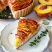 Auf einem weißen Teller liegt eine mit frischen Kräutern garnierte Scheibe Pfirsichtarte. Die ganze Torte und die halbierten Pfirsiche stehen im Hintergrund und zeigen die glänzenden, karamellisierten Pfirsichspalten - perfekt für einen sommerlichen Sommerkuchengenuss.