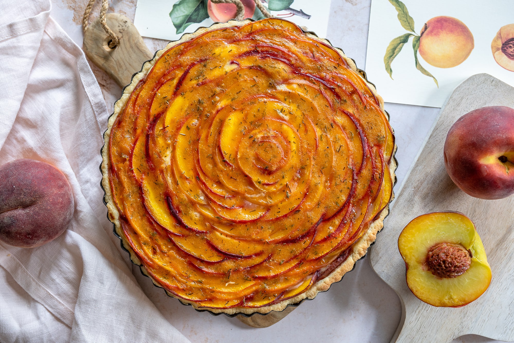 Eine Pfirsichtarte mit spiralförmig angeordneten, dünn geschnittenen Pfirsichen liegt auf einem Holzbrett, umgeben von frischen Pfirsichen, einer Pfirsichhälfte und botanischen Illustrationen - perfekt für jeden Sommerkuchenaufstrich.