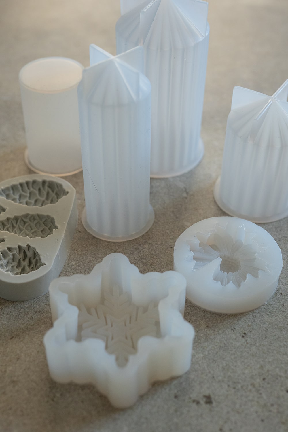 Mehrere weiße Silikonformen in verschiedenen Formen, darunter ein Stern, eine Blume und ein Tannenzapfen, sind auf einer grauen Fläche angeordnet. Die Formen haben detaillierte Muster und werden zum Basteln oder Backen verwendet.