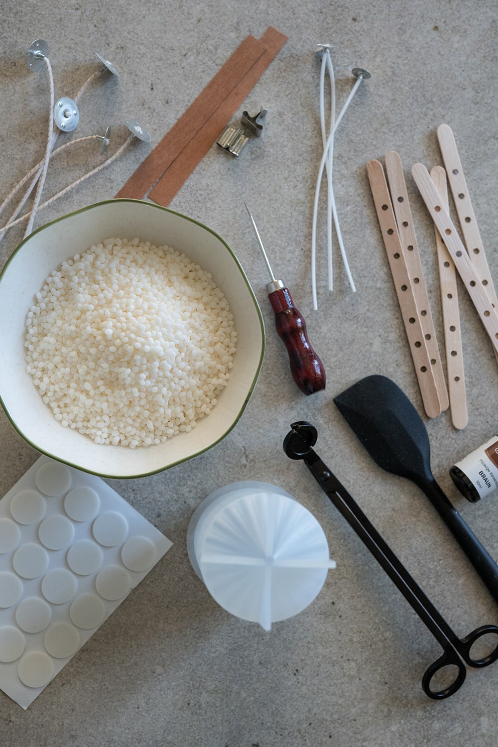 Eine flache Lage von Kerzenherstellungszubehör, darunter Wachspellets in einer Schale, Dochte, Holzstäbchen, ein Schraubenzieher, Plastikbänder, ein Spatel, eine Tropfflasche, Klebepunkte und ein Messlöffel auf einer beigen Oberfläche.