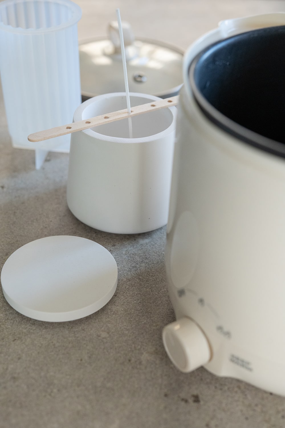 Ein Kerzenherstellungsset mit einem weißen Wachsschmelzer, einem weißen Behälter mit einem Docht, der von einem Holzstab aufrecht gehalten wird, einem Deckel und einer Silikonform, die alle auf einer grauen Fläche stehen.