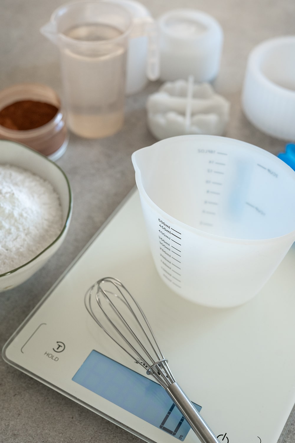 Eine digitale Küchenwaage mit einem Schneebesen und einem leeren Messbecher darauf, umgeben von einer Schale mit weißem Pulver, einem Behälter mit Kakao, einer Tasse mit Wasser und anderen Küchengeräten auf einer Arbeitsplatte.