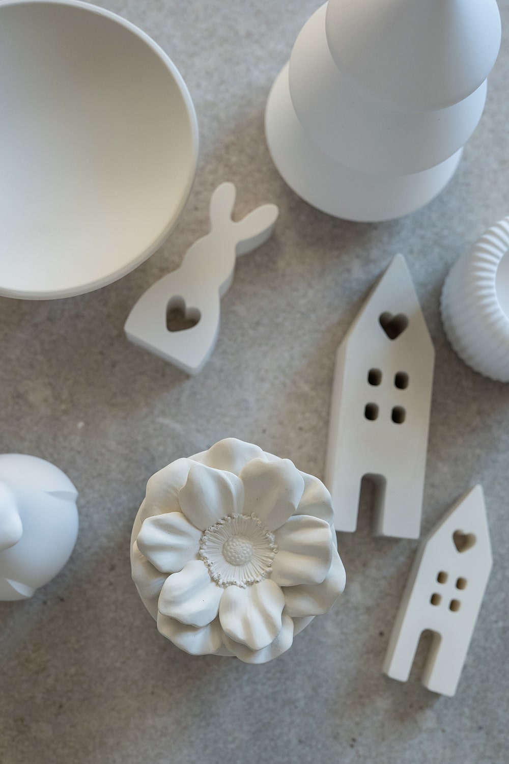 Weiße keramische Dekorationsobjekte, darunter eine Blume, zwei Häuser, ein Hase mit Herzausschnitt, eine Schale und abstrakte Formen, sind auf einer hellgrauen, strukturierten Oberfläche angeordnet.