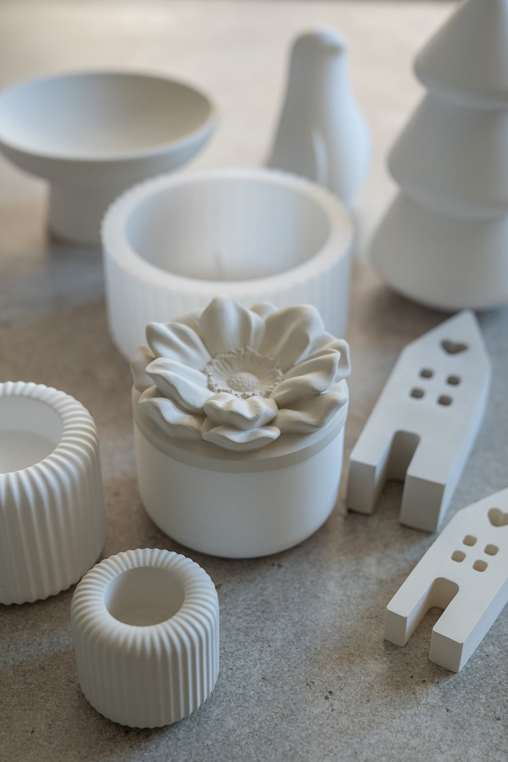 Eine Sammlung von weißen Keramikobjekten, darunter gerippte Schalen, ein Behälter mit blumenförmigem Deckel, minimalistische Haus- und Baumformen sowie glatte, skulpturale Formen, die auf einer neutralen Oberfläche angeordnet sind.