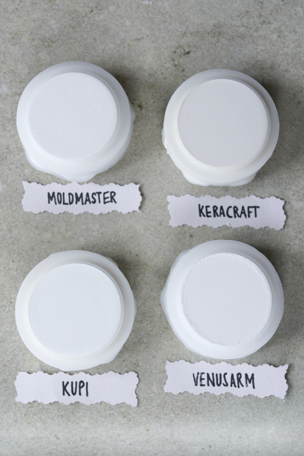 Vier weiße runde Deckel mit unterschiedlich glatten Oberseiten sind in einem Raster auf einer hellen Fläche angeordnet. Jeder ist mit handgeschriebenen Namen auf Papier beschriftet, perfekt für einen Reliefpulver Vergleich oder um Tipps für Reliefpulver zu entdecken.