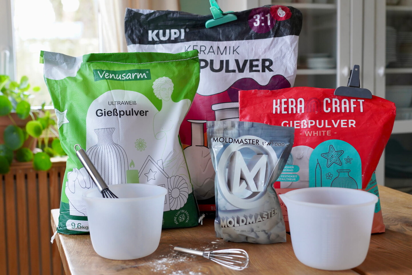 Tüten mit Gießpulvern und Bausätzen, Mischschüsseln, ein kleiner Schneebesen und Pulver auf einem Holztisch - mit Pflanzen und einem Schrank im Hintergrund. Die farbenfrohe Verpackung deutet auf Bestes Reliefpulver für kreative Bastelarbeiten oder Ihren nächsten Reliefpulver Vergleich hin.