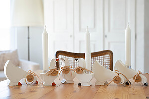 Ida & Emil - handgefertigte Gänse-Kerzenhalter im skandinavischen Stil mit Namensschildern und Schleifen stehen auf einem Holztisch neben zwei weißen Kerzen in einem hellen, gemütlichen Raum.