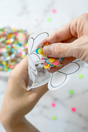 Eine Hand füllt einen mit Herzumrissen verzierten Papierkegel mit buntem Konfetti. Im Hintergrund steht verschwommen auf einer Marmorfläche eine Schale mit weiterem Konfetti.
